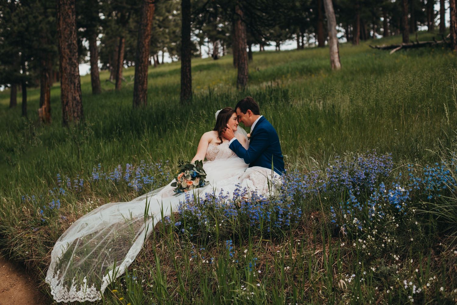 Newlyweds share intimate moment sitting in wildflower field with long veil flowing behind.