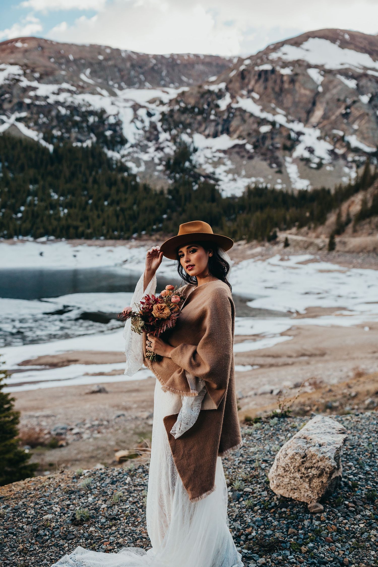 A wedding photo in the snowy mountains with a brown coat over a white dress and autumn bouquet.