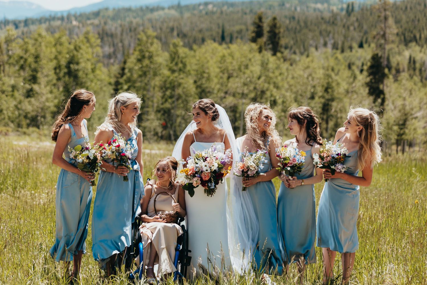 Wedding party in light blue dresses poses together in a sunny mountain meadow with flowers.