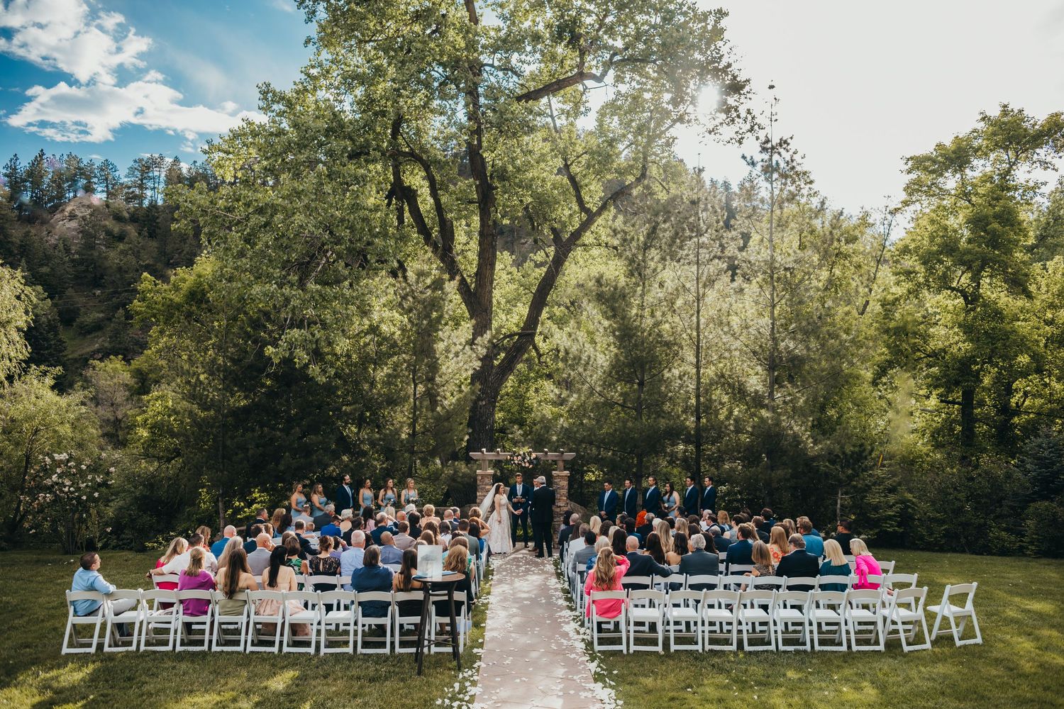 Outdoor wedding ceremony setup with white chairs in wooded setting under bright blue sky.