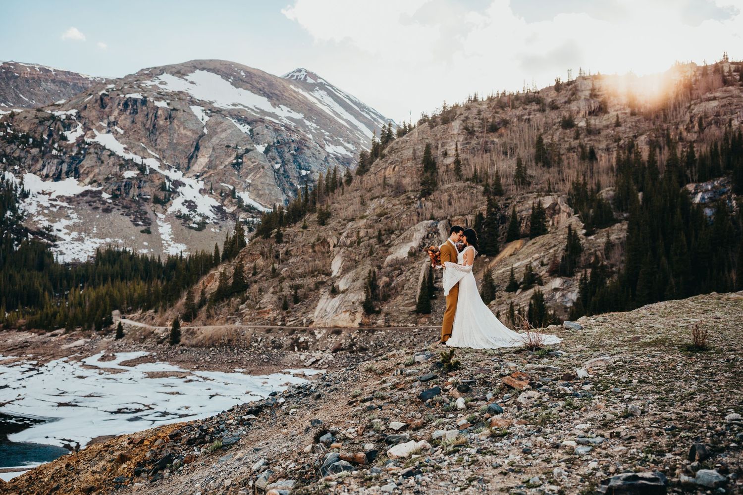 Wedding couple celebrates on mountain cliff edge with dramatic landscape and frozen lake below.