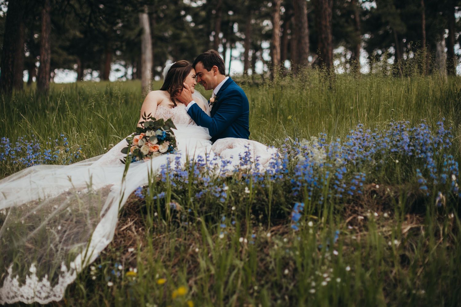 Newlyweds embrace in field of blue wildflowers with pine trees in background.