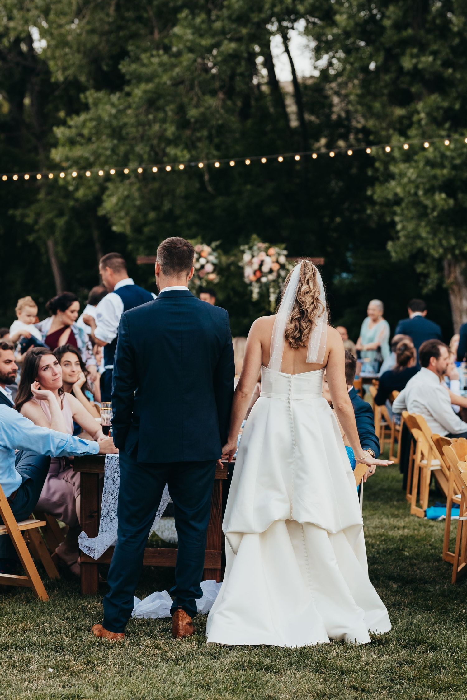 A couple stands at an outdoor wedding reception with string lights and wooden chairs under trees at dusk.