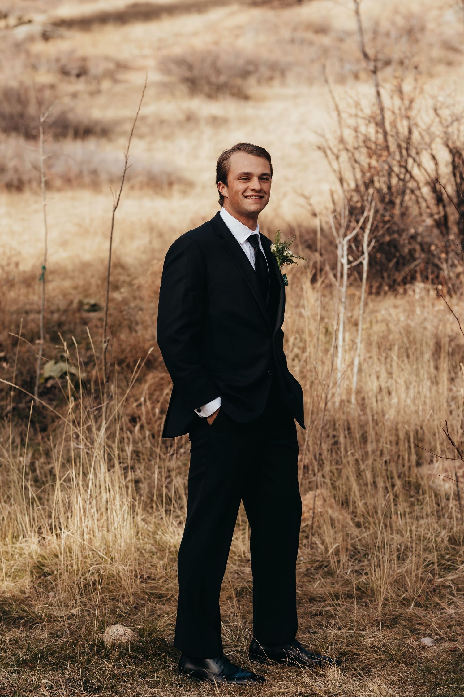 Groom in classic black suit poses against autumn landscape backdrop.