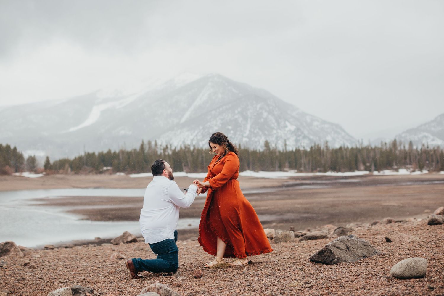 Marriage proposal scene with mountain lake and snow-capped peaks in background.