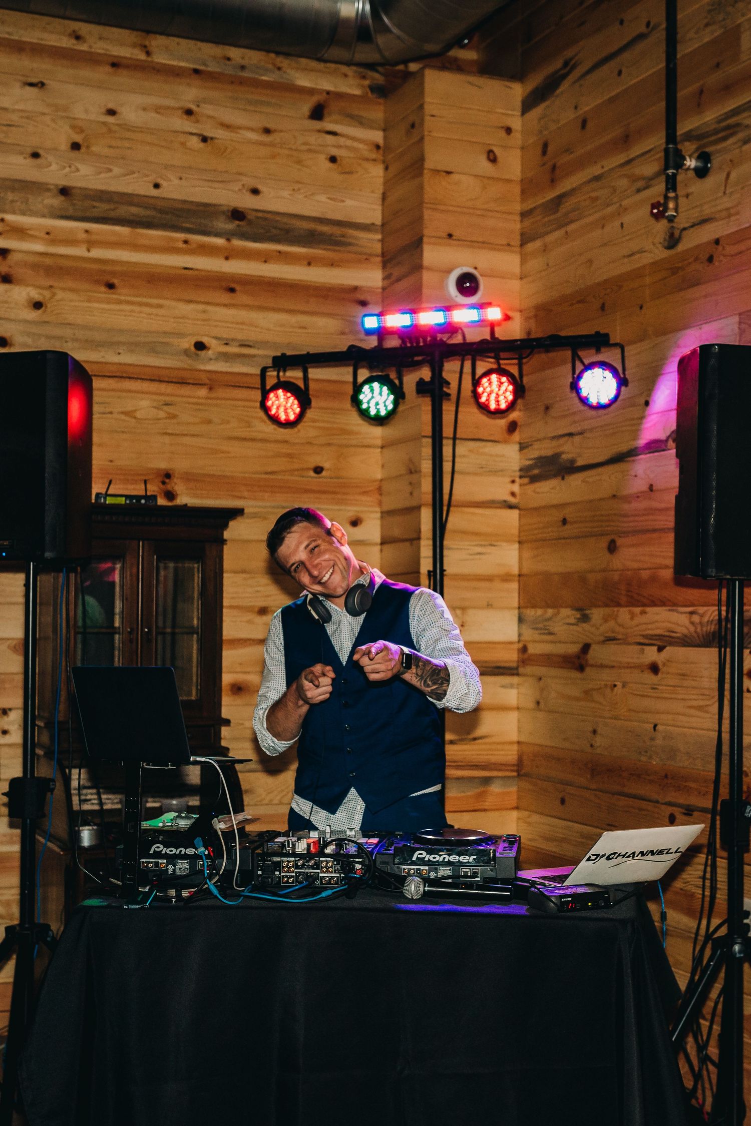 A DJ performs enthusiastically at a wedding reception with colorful stage lights in a wooden venue.
