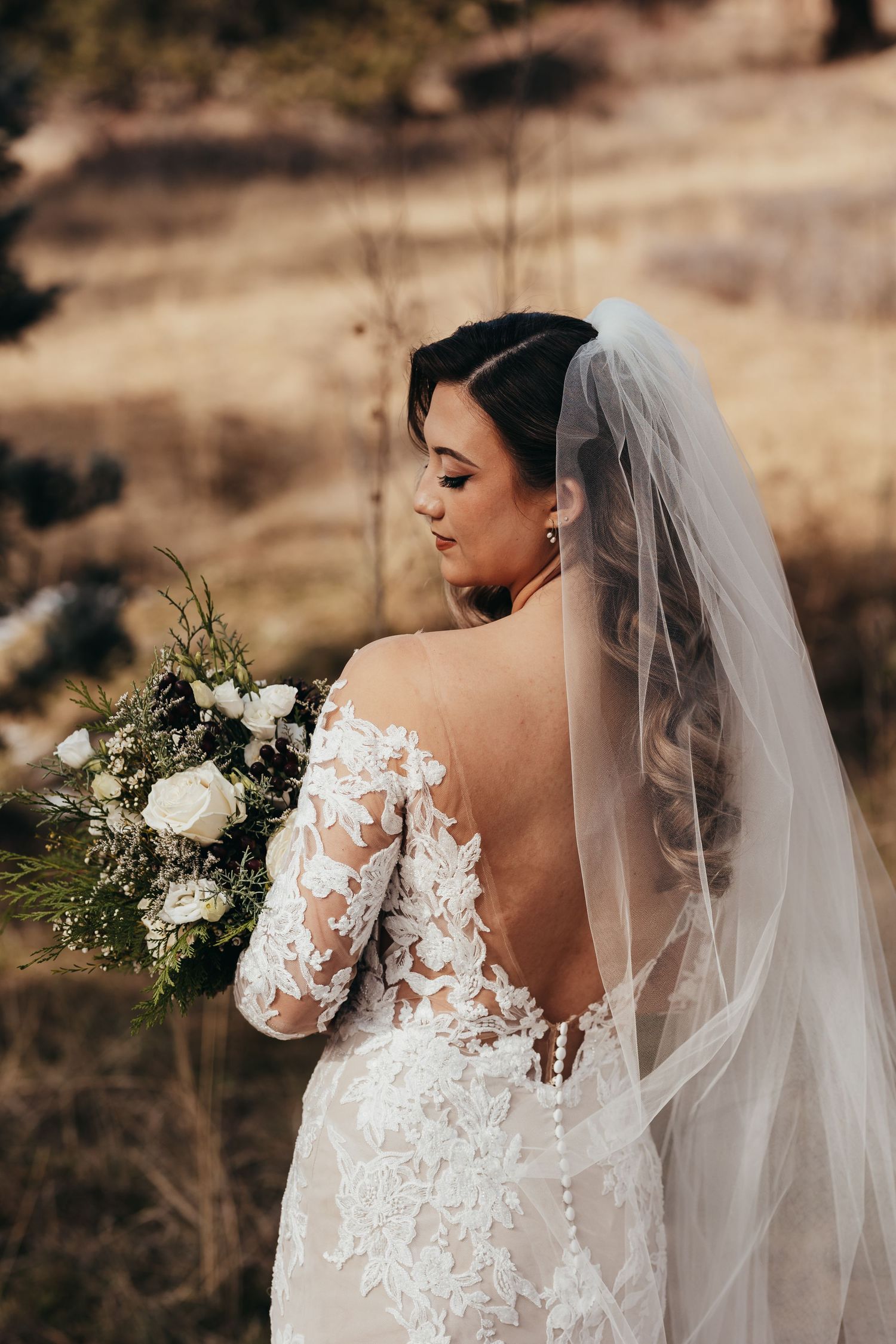 Back view of bride in lace dress with flowing veil holding white floral bouquet.