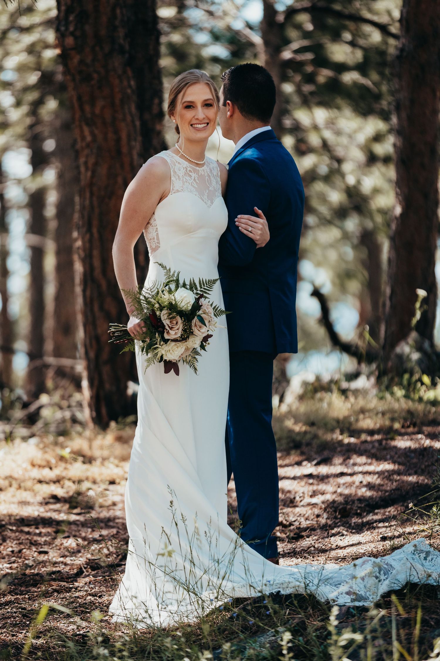 Romantic forest wedding portrait with couple in white dress and navy suit among tall pine trees.