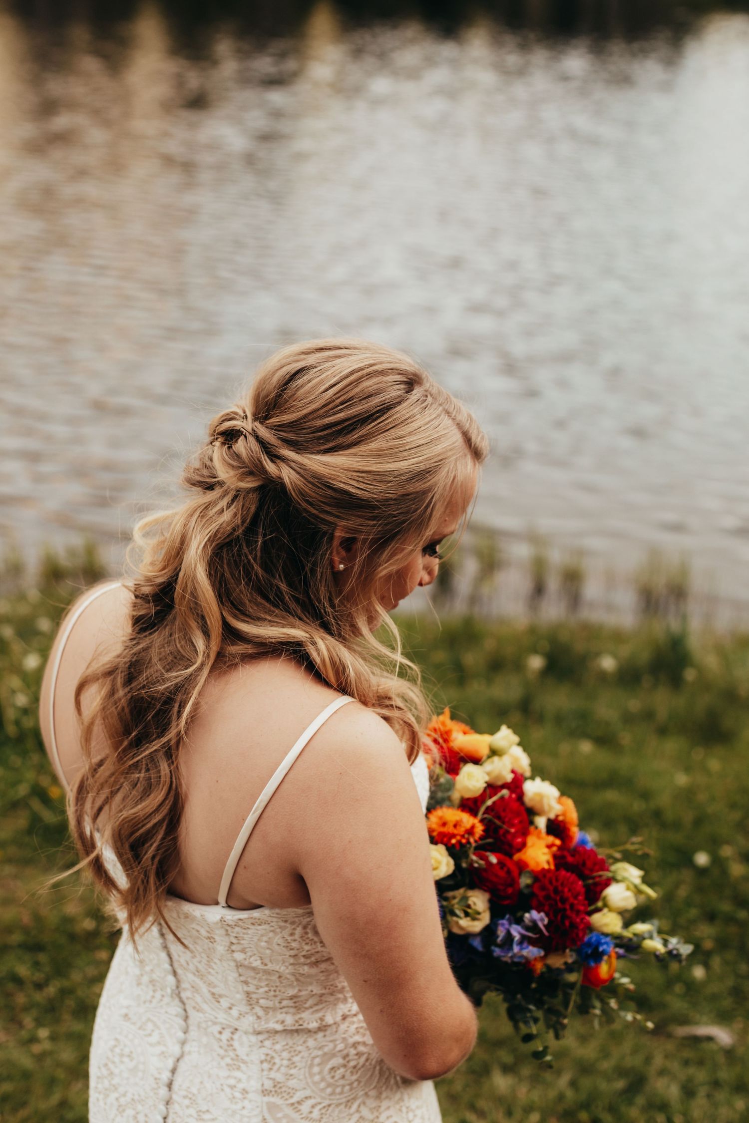 A bride in a lace dress holds a vibrant fall-colored bouquet by a lake.