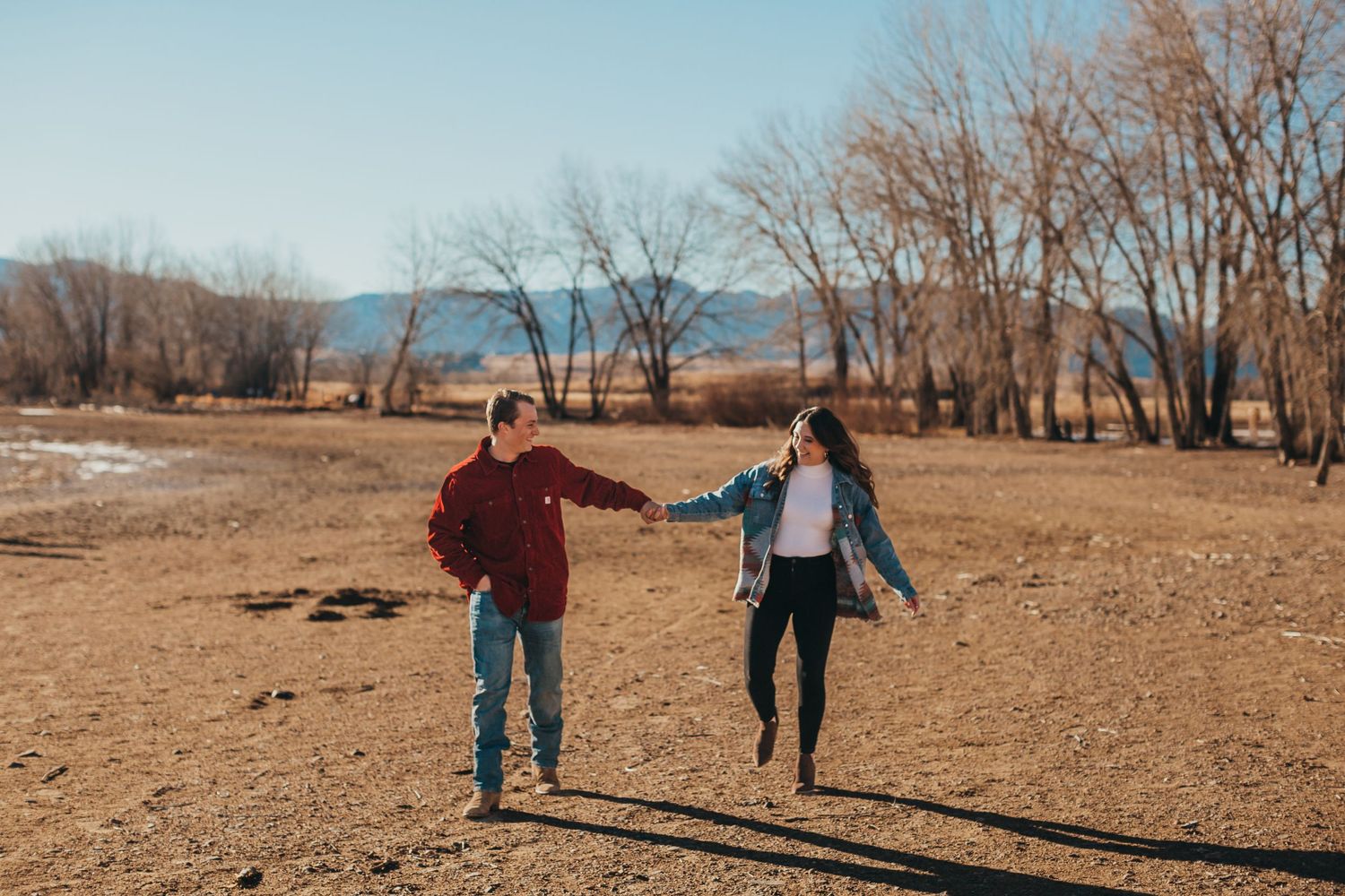 Couple holds hands while walking through an open field with mountains in background.