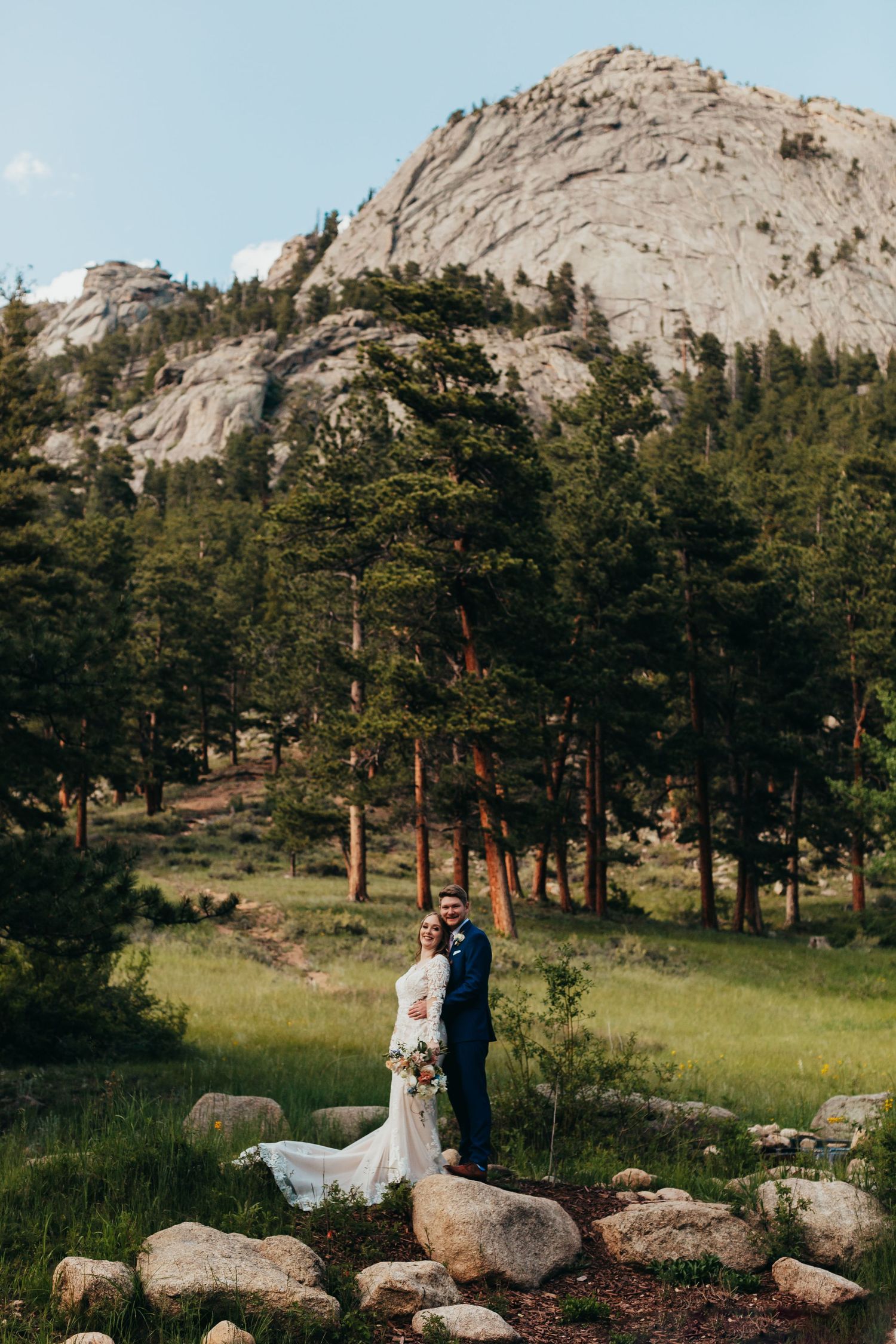 A wedding couple poses in front of a majestic rocky mountain backdrop with tall pine trees in a scenic natural setting.