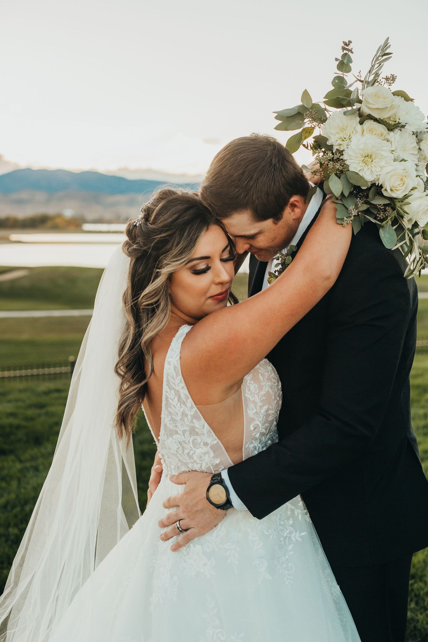 A romantic wedding couple embraces outdoors with mountains in the background as the bride holds a white floral bouquet.
