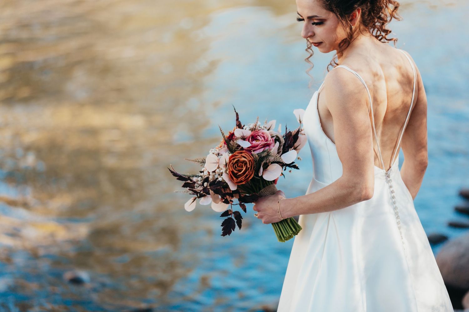 Side view of bride in backless dress holding burgundy bouquet against water backdrop.