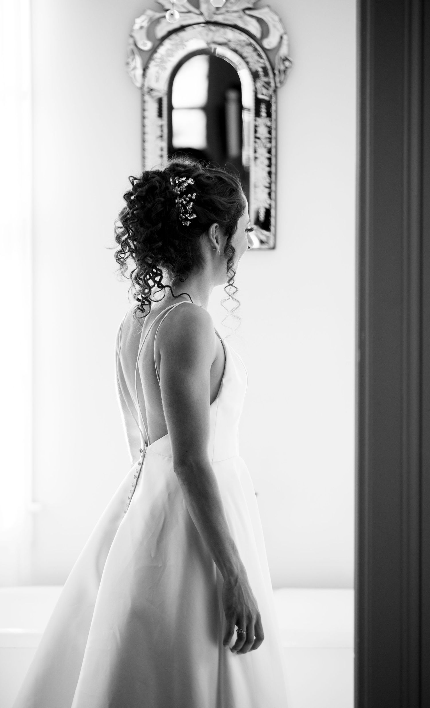 Black and white photo of bride in elegant dress looking at ornate mirror.