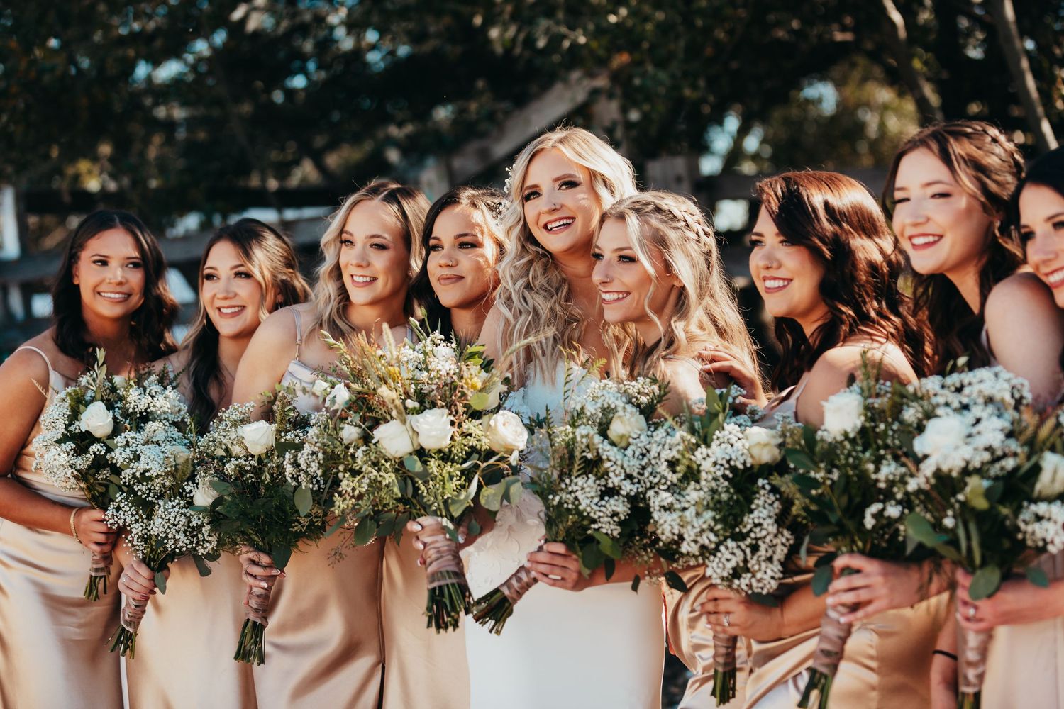 Bridesmaids in champagne dresses holding bouquets pose together outdoors.