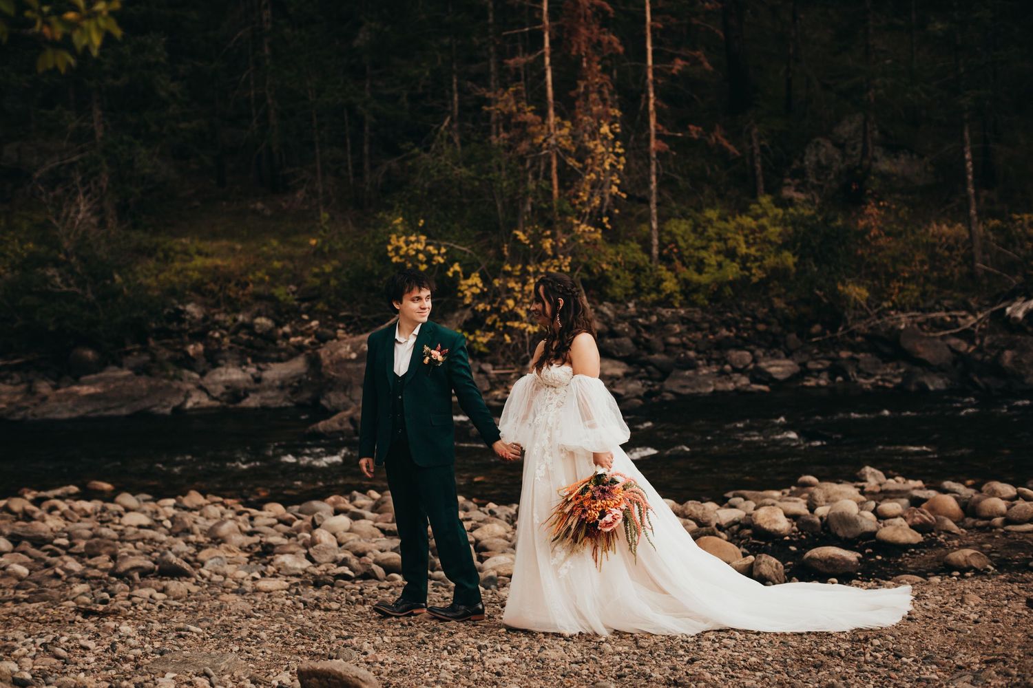 Wedding couple poses on rocky riverbank surrounded by autumn trees.