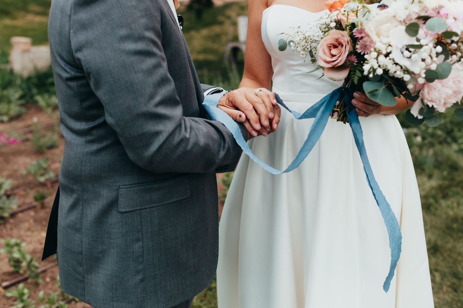 Close up of couple's hands during wedding ceremony with bride holding floral bouquet with blue ribbon.