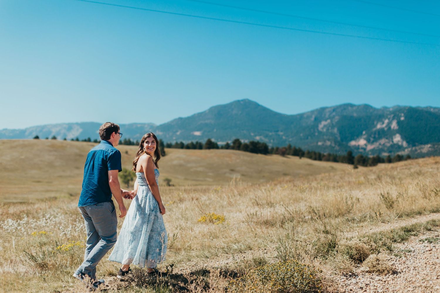 Couple dancing in mountain meadow with blue sky and rolling hills backdrop.