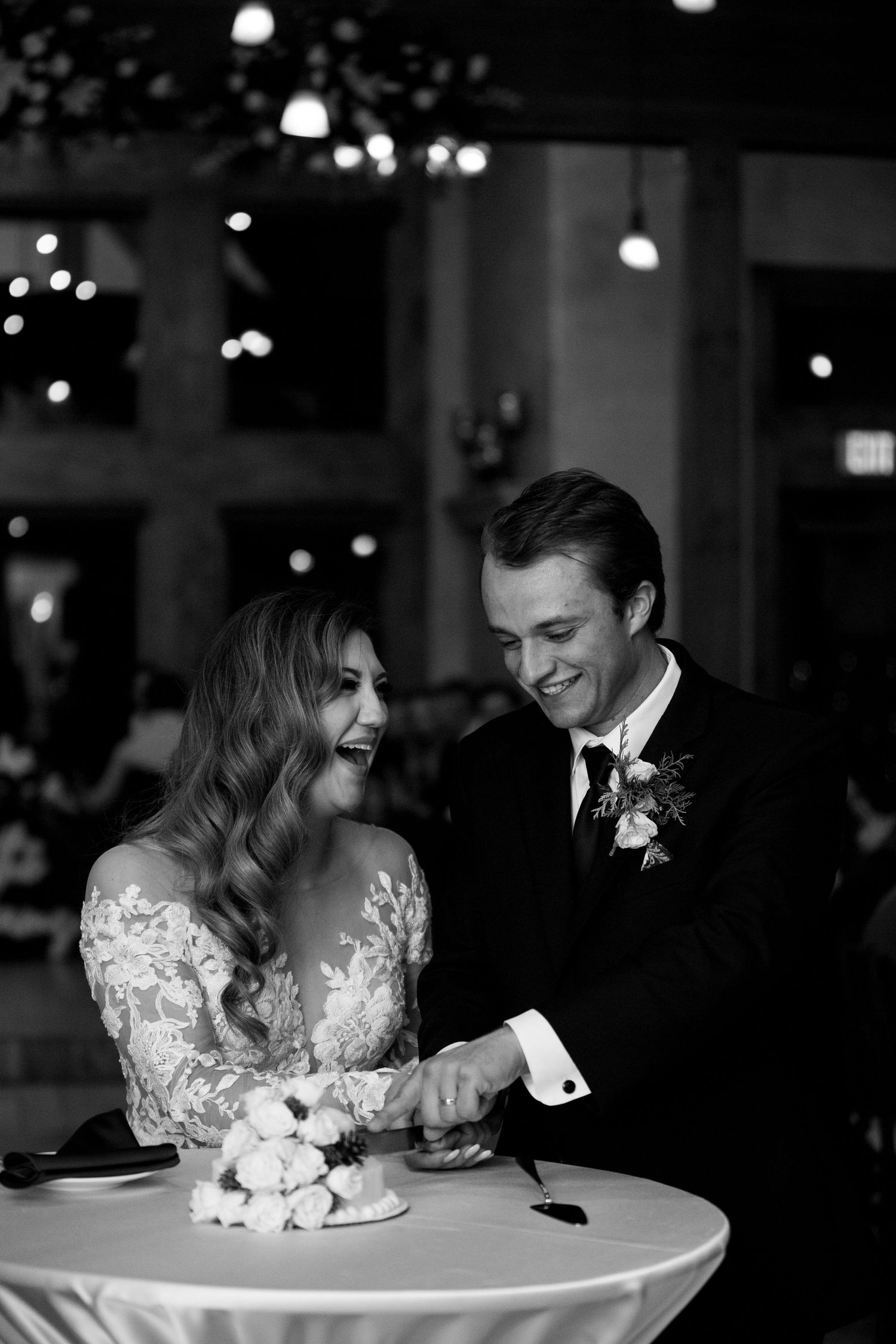 Black and white candid shot of couple sharing laughter while cutting their wedding cake.