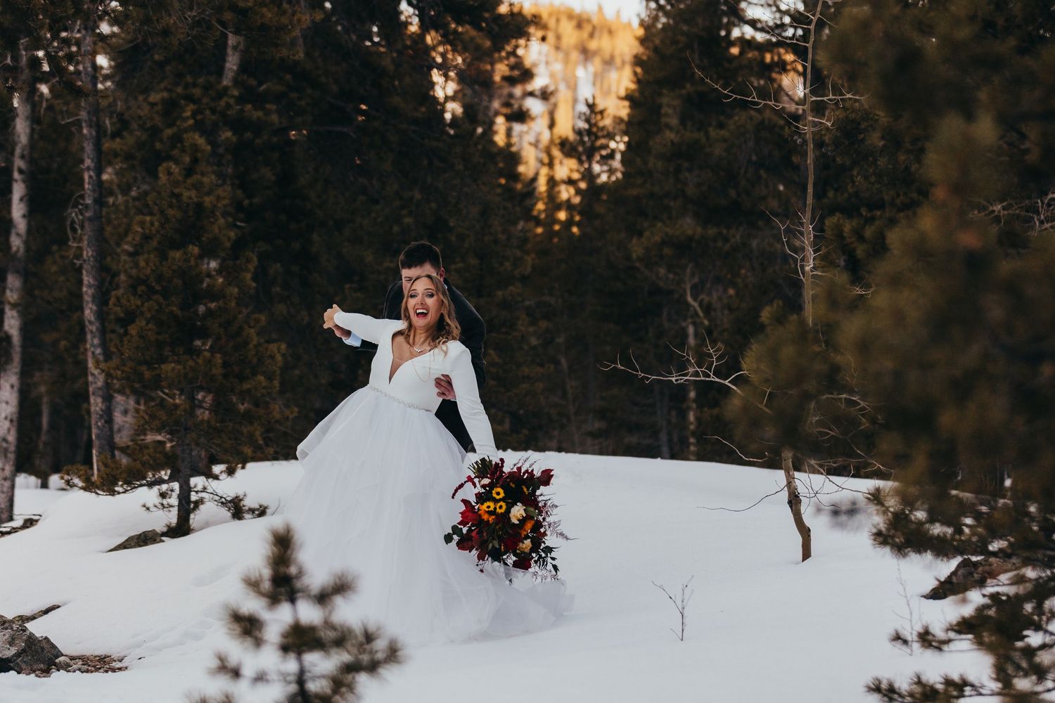 A bride in a white wedding dress walking through a snowy forest holding a colorful bouquet.