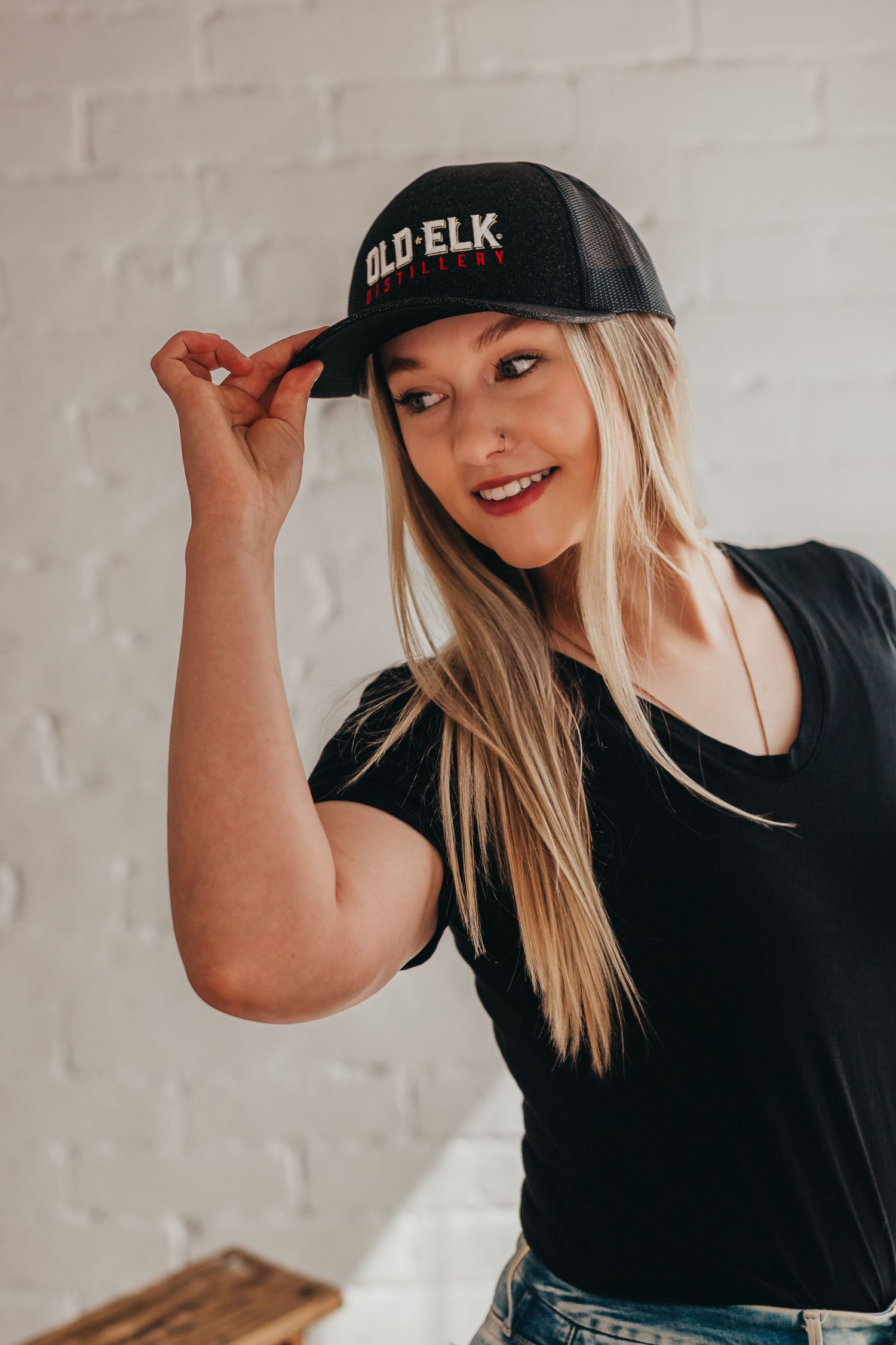 Portrait of someone wearing a black Old Elk baseball cap and black t-shirt against a white brick wall.
