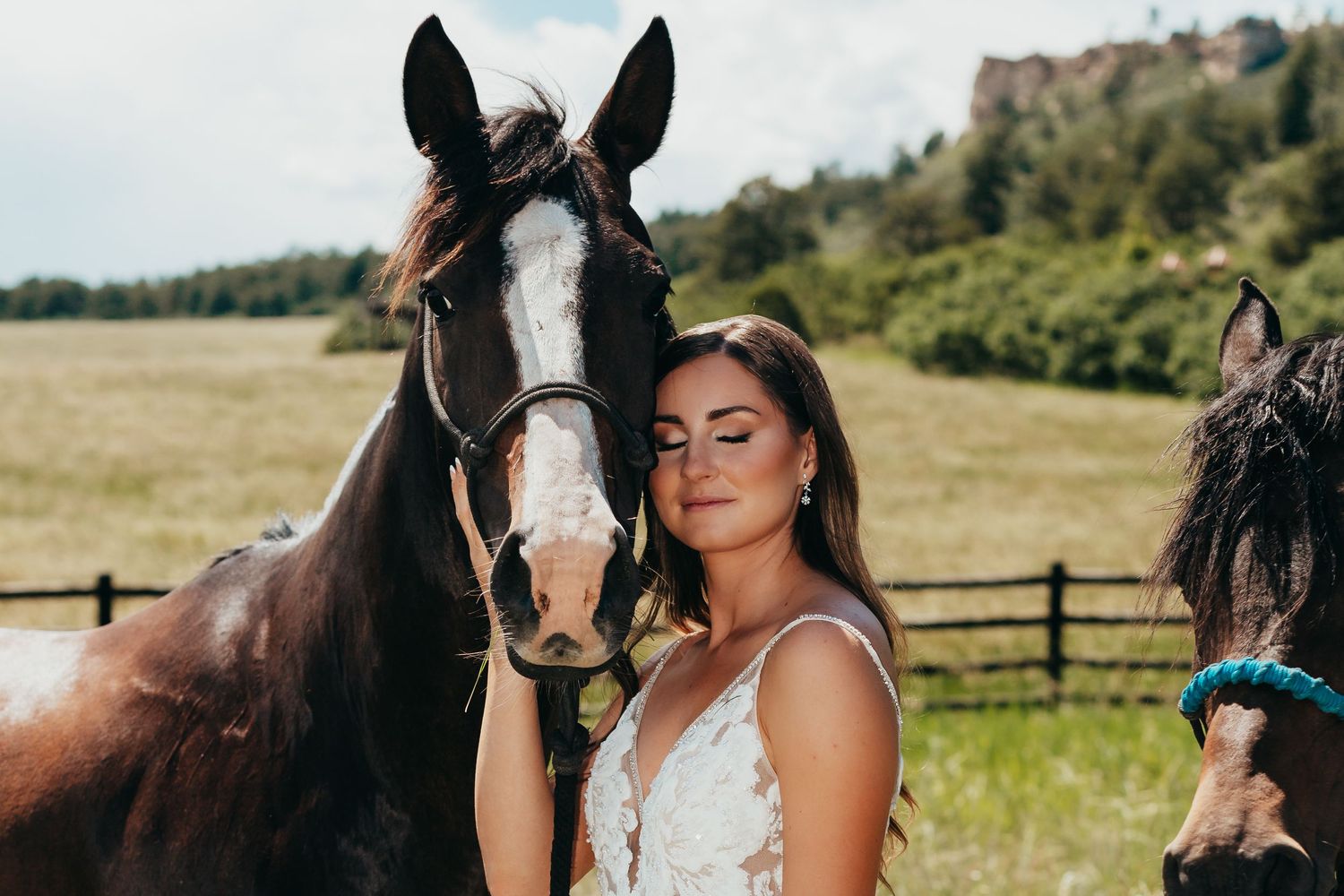 A brown horse with white blaze stands next to someone in a white lace dress against mountain backdrop.