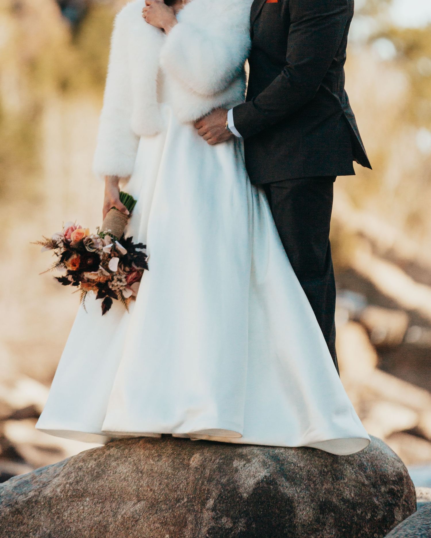 Couple stands on rock with bride holding autumn bouquet.