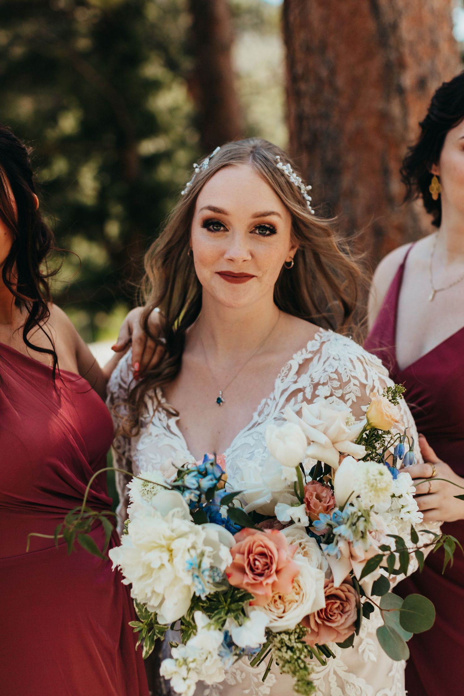 A bride in a lace wedding dress holds a colorful bouquet of roses and delphinium flowers.