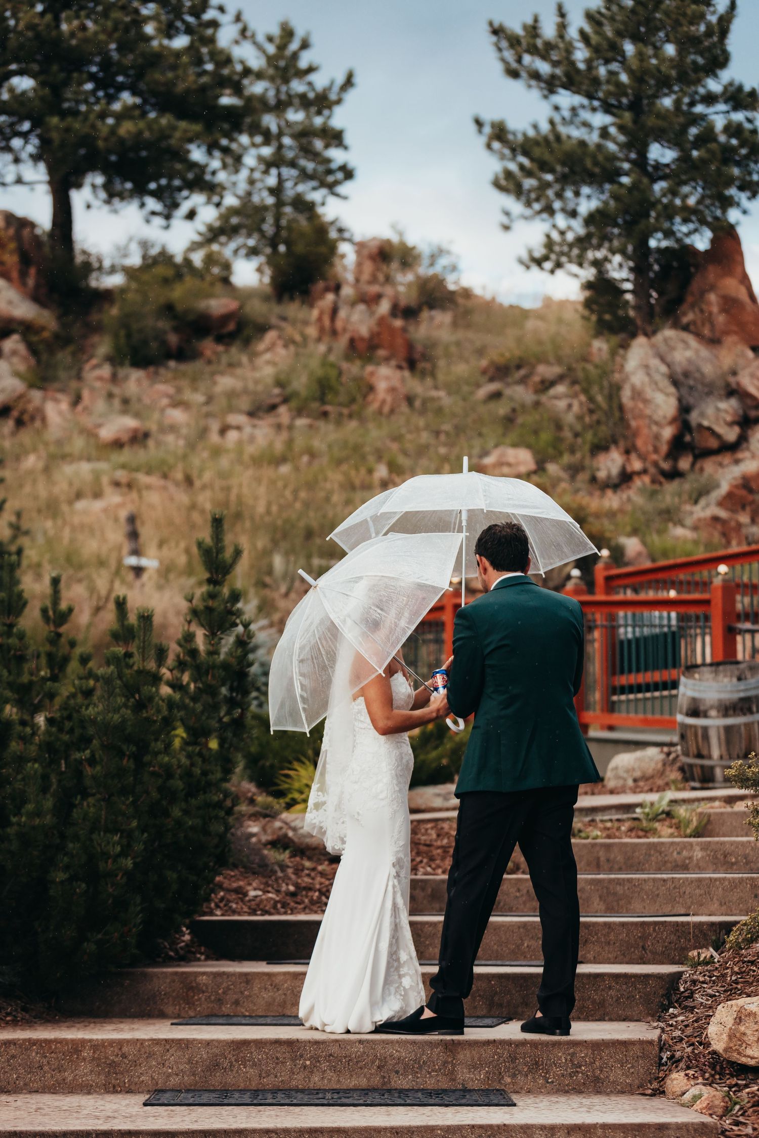 Wedding couple shares an intimate moment under clear umbrellas on stone steps surrounded by mountain scenery.