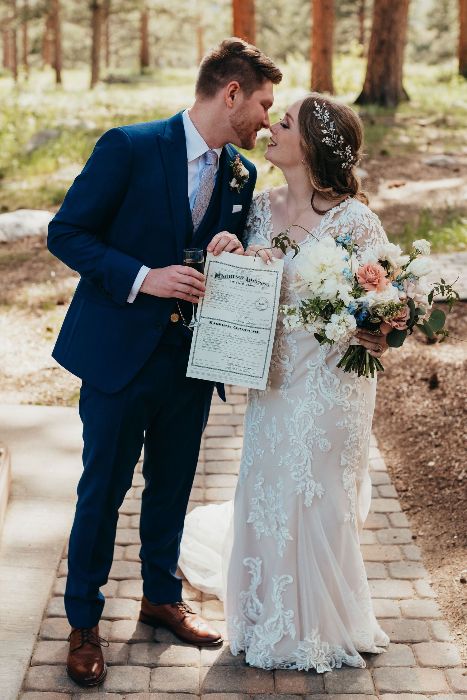 A couple holds their marriage certificate while standing on a stone pathway surrounded by pine trees.