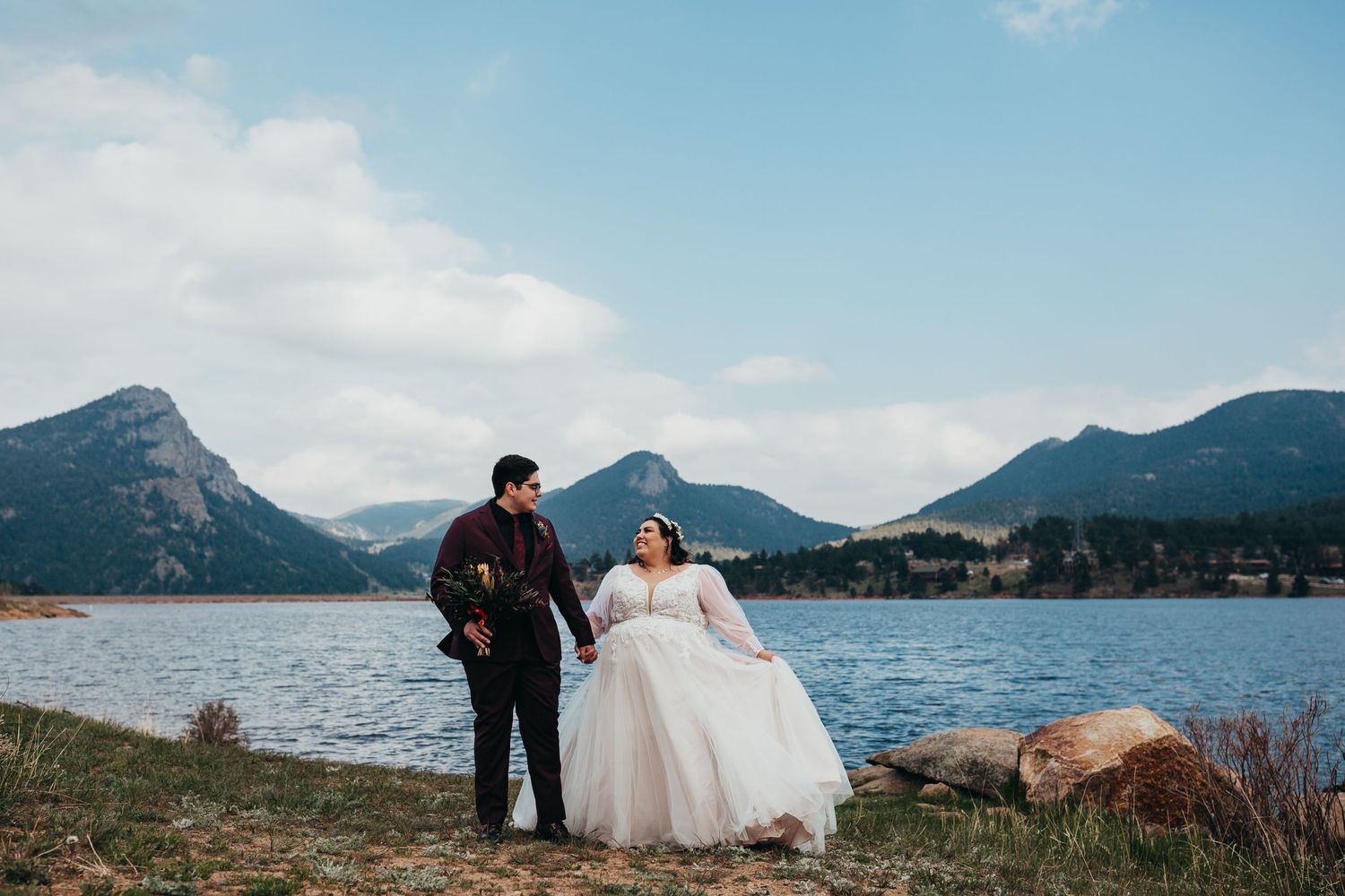 Wedding couple standing by mountain lake with dramatic landscape backdrop.