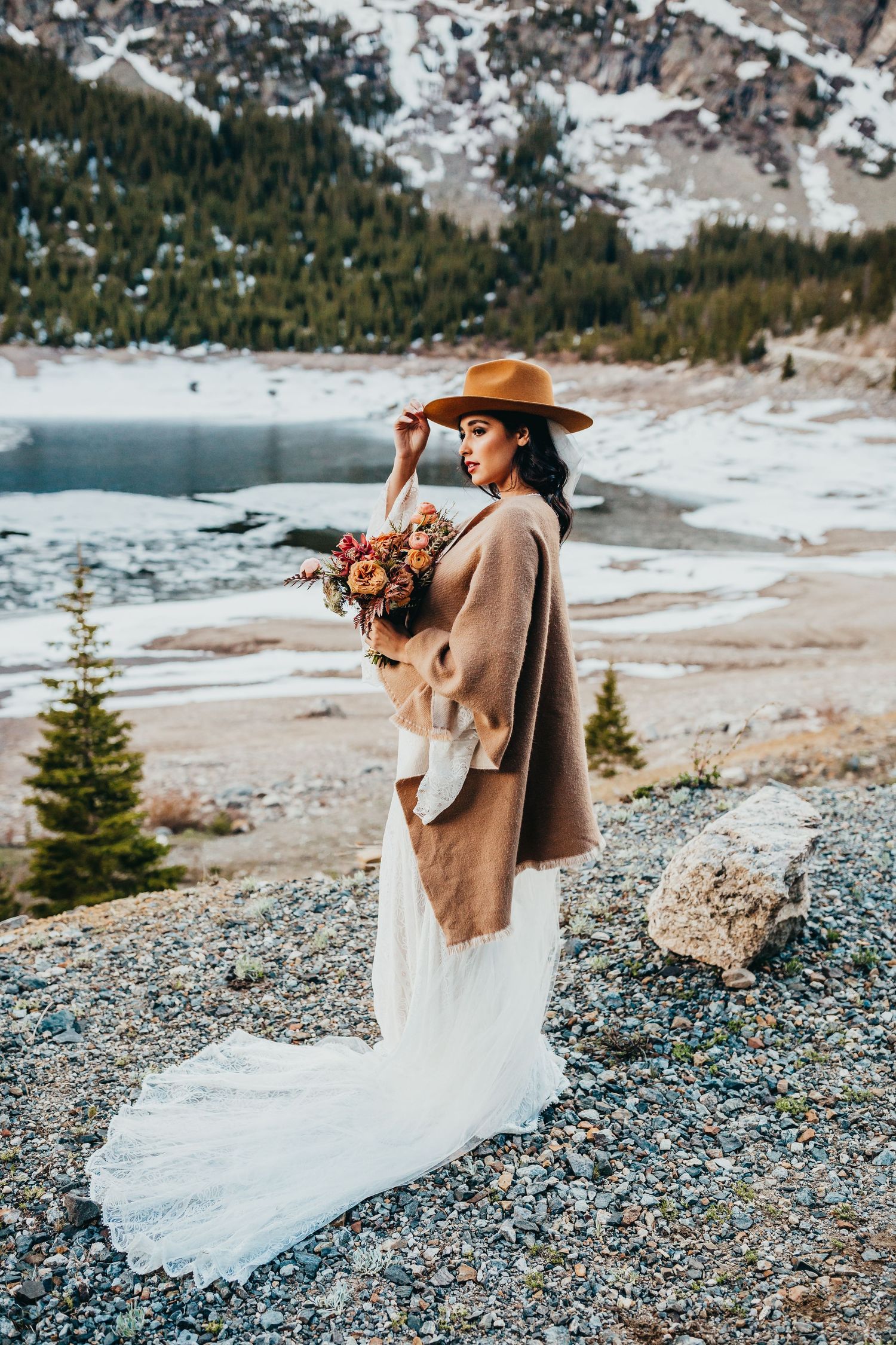 A winter bridal portrait by a mountain lake featuring a long white dress and camel coat.