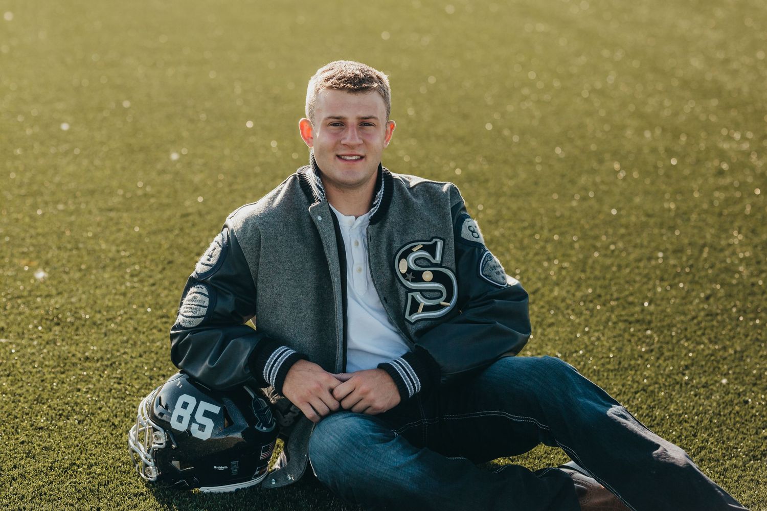 Senior photo of student in varsity jacket posing with football helmet on sunny field.