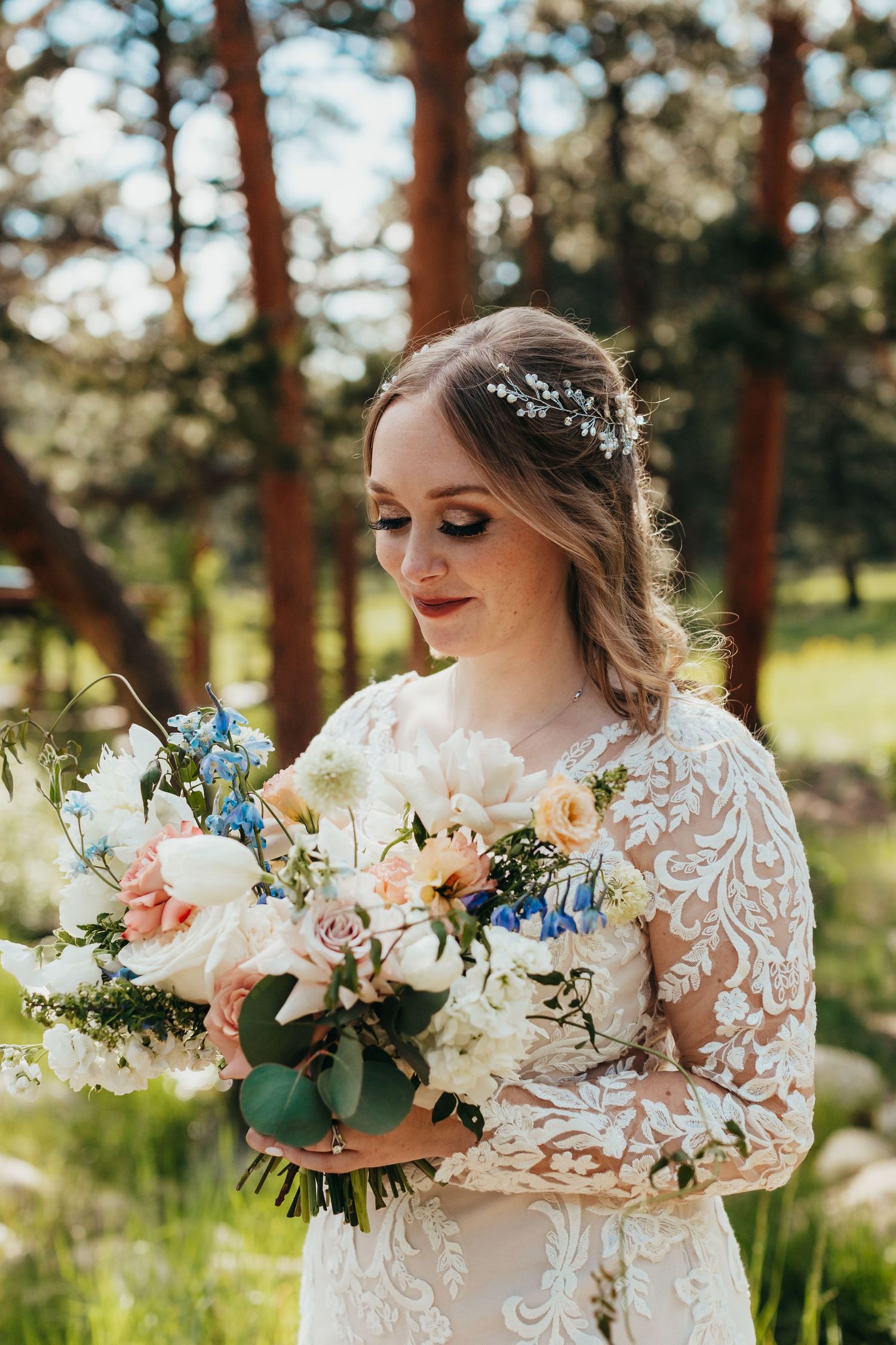 A bride holds a stunning bouquet with white and blue flowers against a sunlit forest backdrop.