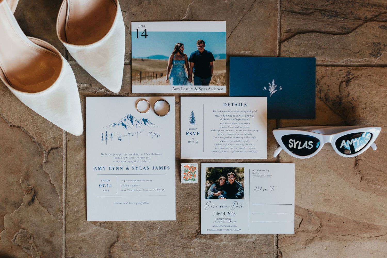 Wedding invitation suite with white heels and sunglasses on a stone surface.
