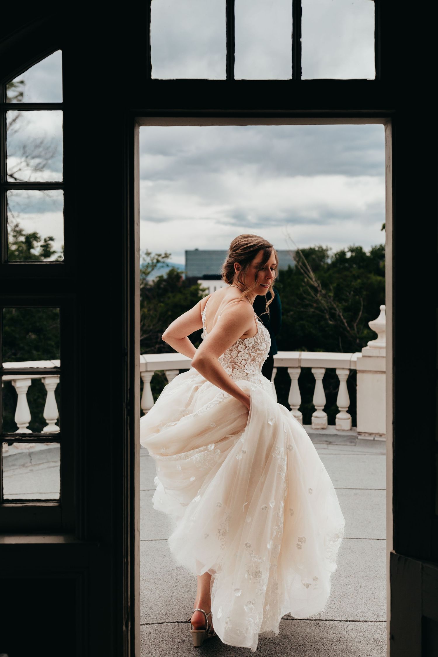 A bride twirls her elegant white dress on a balcony against a cloudy sky backdrop.