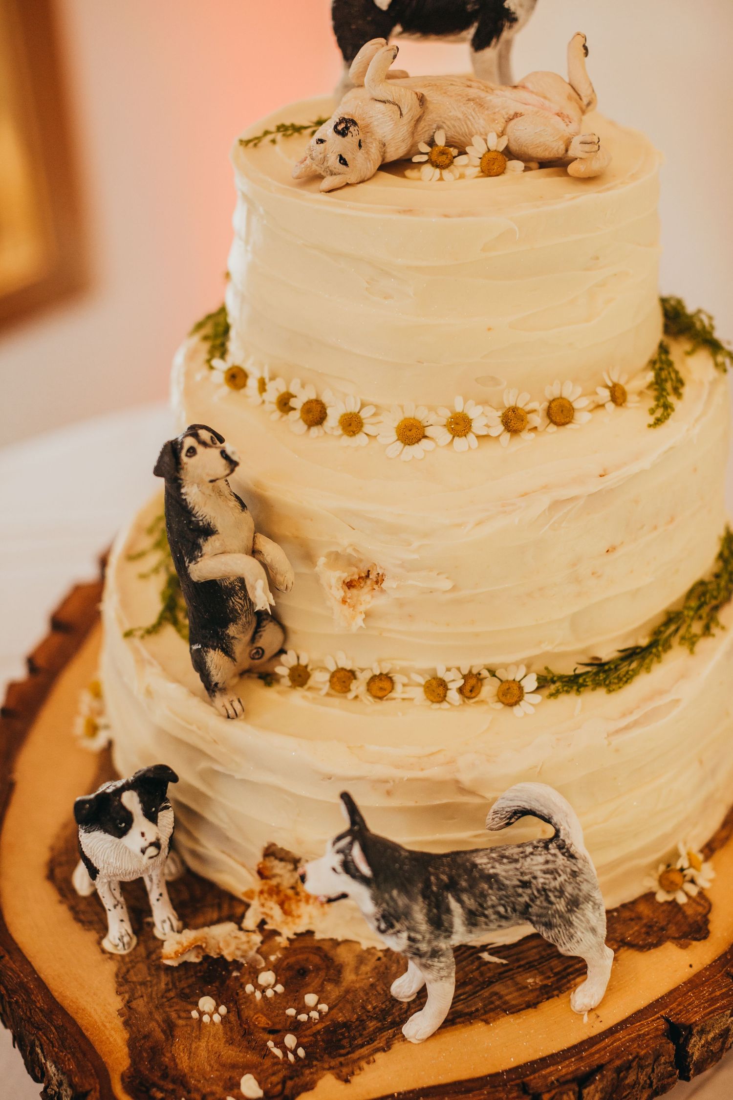 Three-tier white wedding cake decorated with dog figurines and greenery on wooden base.
