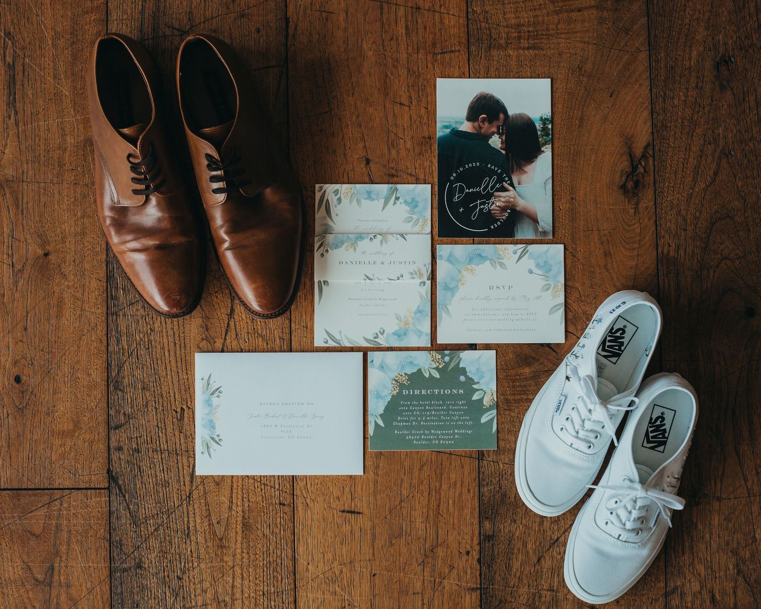 Flat lay of brown dress shoes and white sneakers with wedding stationery and photos on wooden floor.