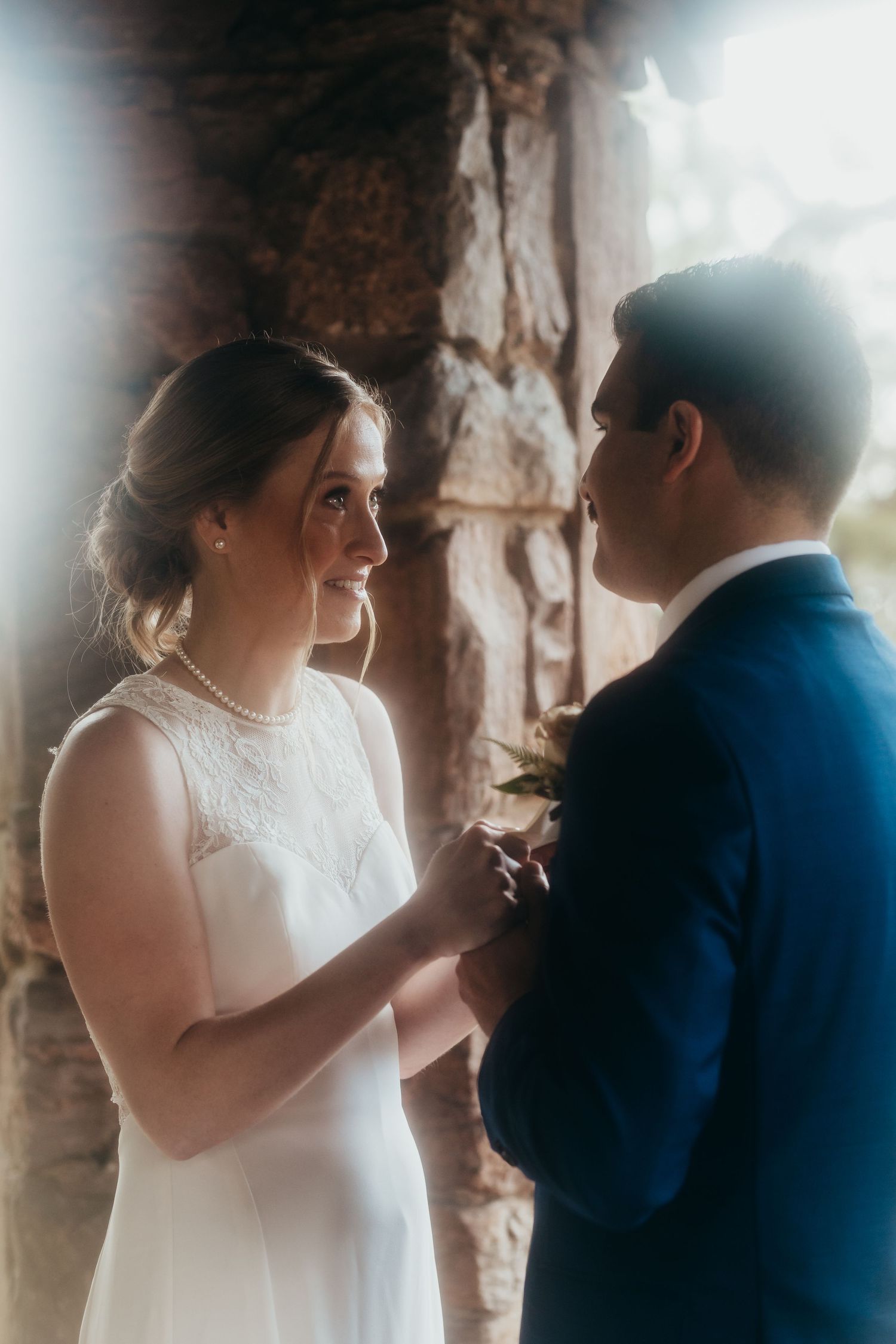 A tender wedding ceremony moment captured in soft lighting against a stone wall backdrop.