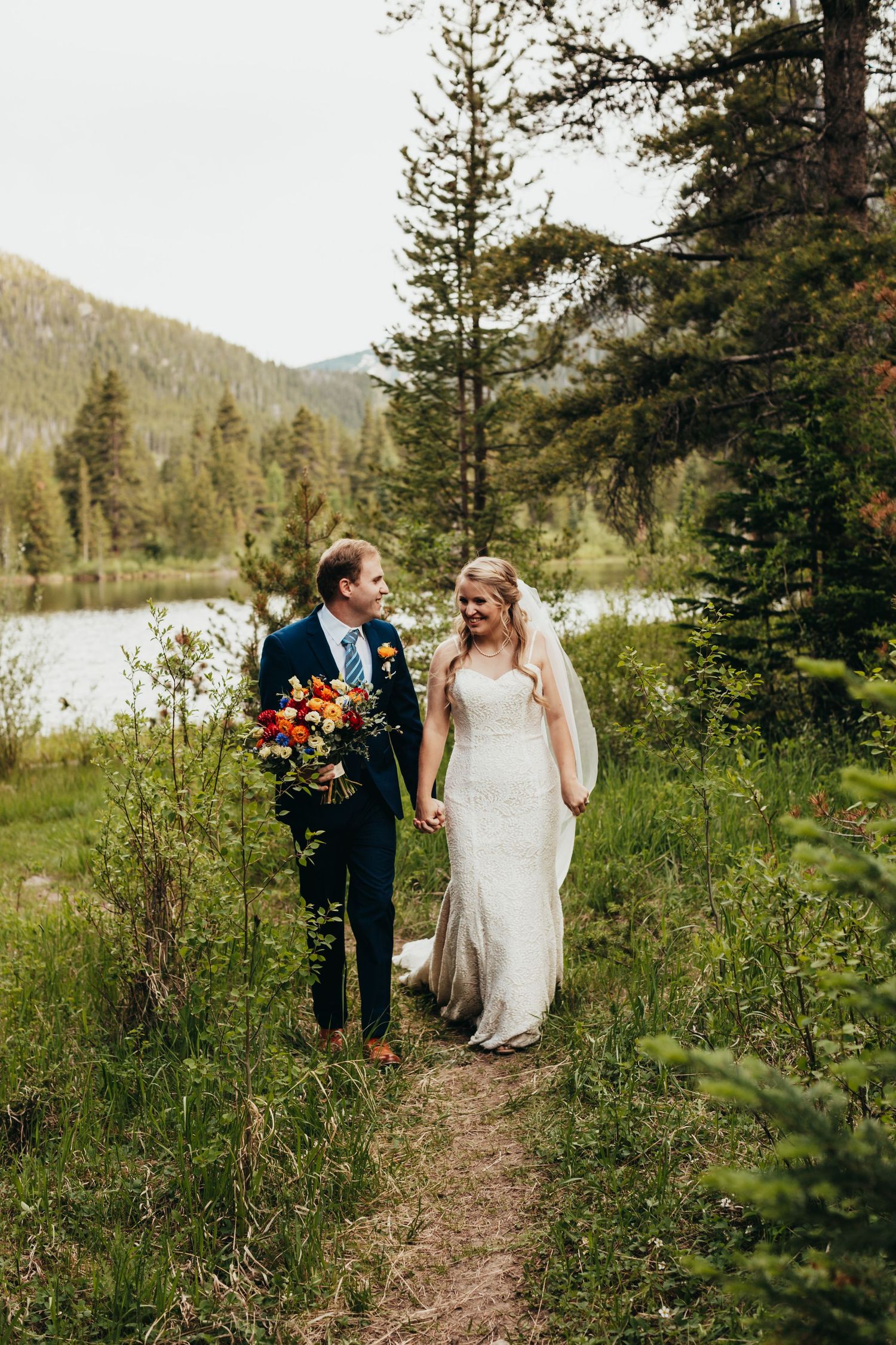 Newlyweds walking on forest trail with vibrant bouquet and mountain scenery.