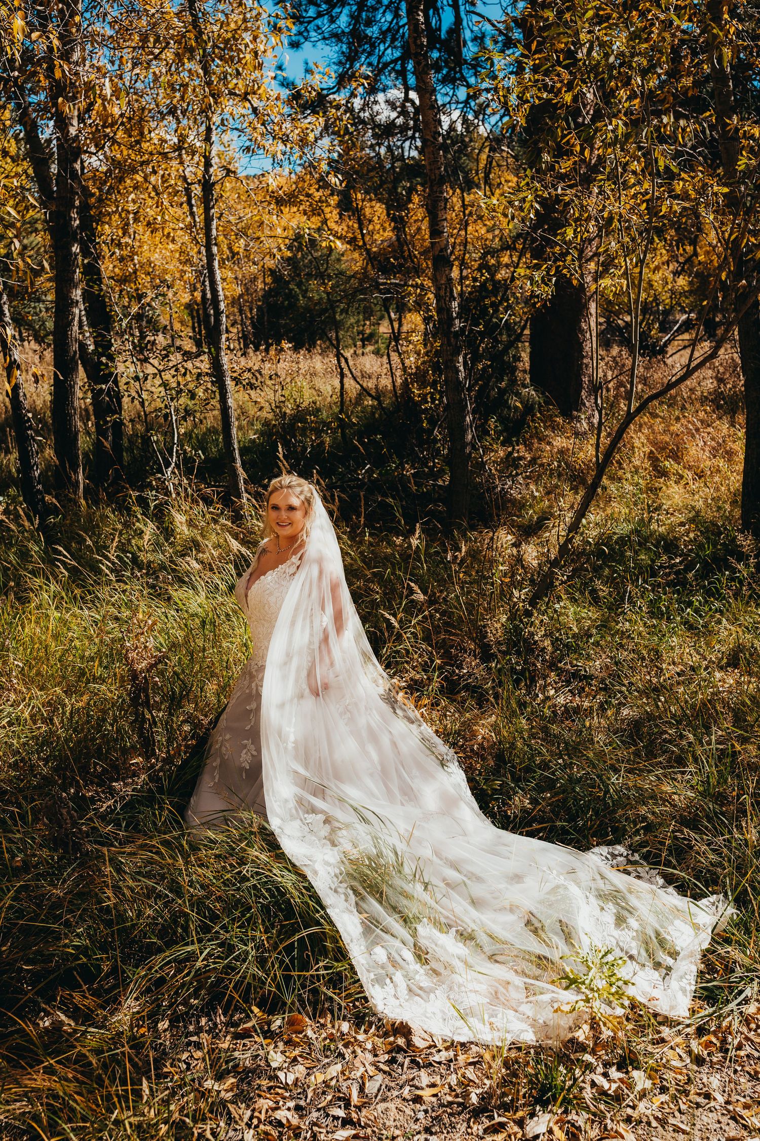 Bride in flowing white dress poses among golden autumn trees.