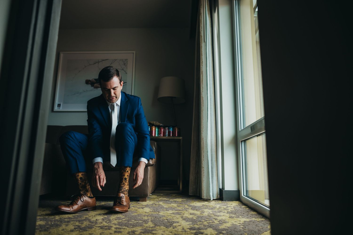 Moody portrait of someone in blue suit adjusting shoes by window with dramatic lighting.
