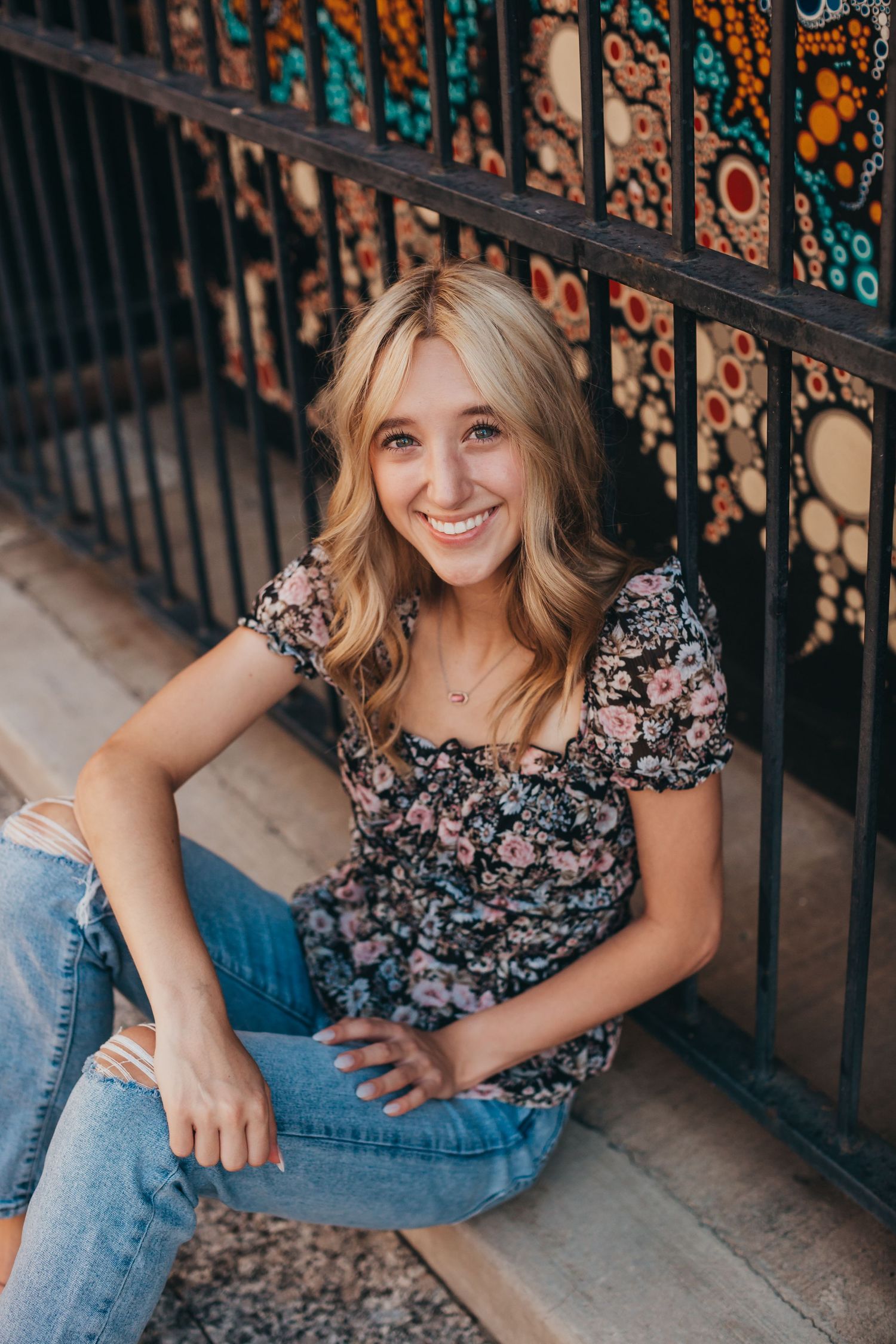 Person seated casually by decorative wrought iron fence wearing floral top and jeans.