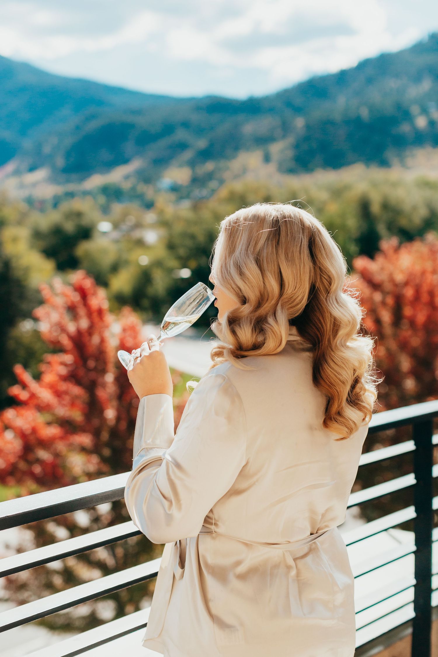 Person enjoying a drink on a balcony overlooking mountains with autumn foliage in the background.