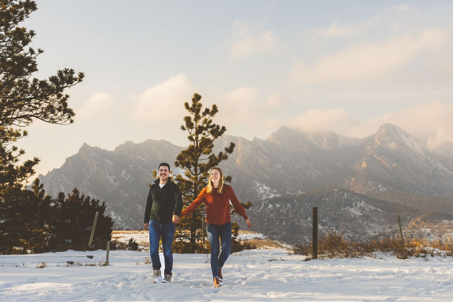 Two people running through snowy mountain terrain during sunset with peaks visible in the background.