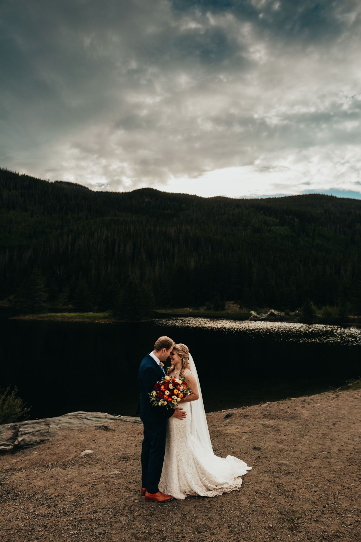 A couple embraces during their lakeside wedding with dramatic storm clouds overhead.