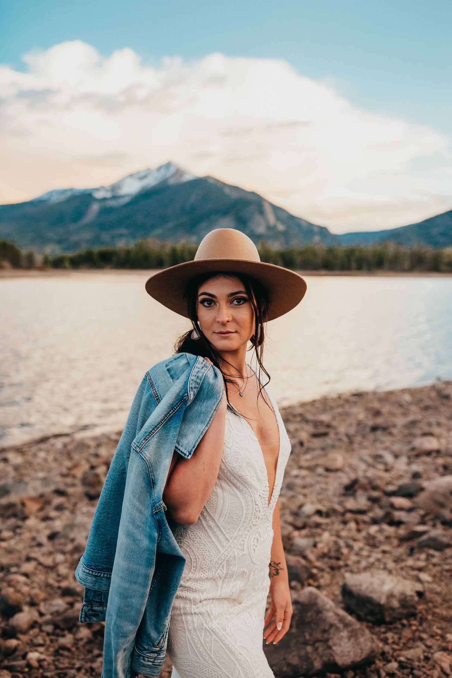 Bride wearing brown hat and denim jacket poses against mountain lake backdrop.