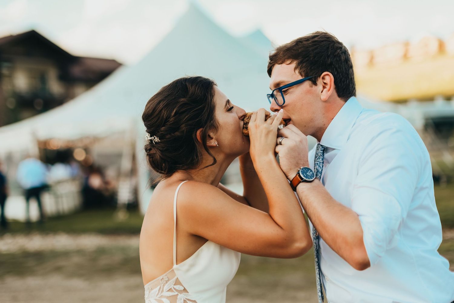 A couple shares a playful moment with a burger at an outdoor reception.