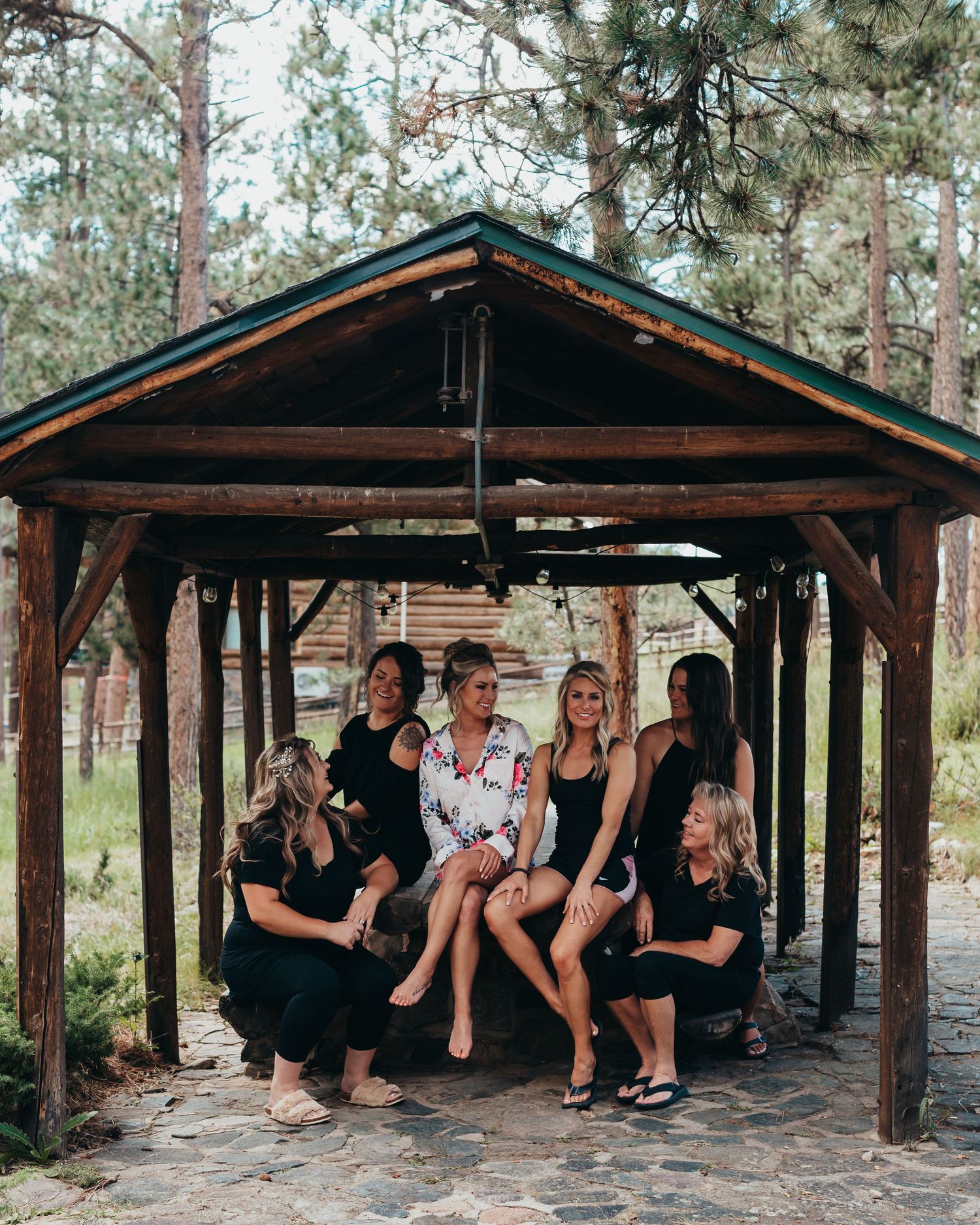 Wedding party posing together under a rustic wooden gazebo in a forest setting.
