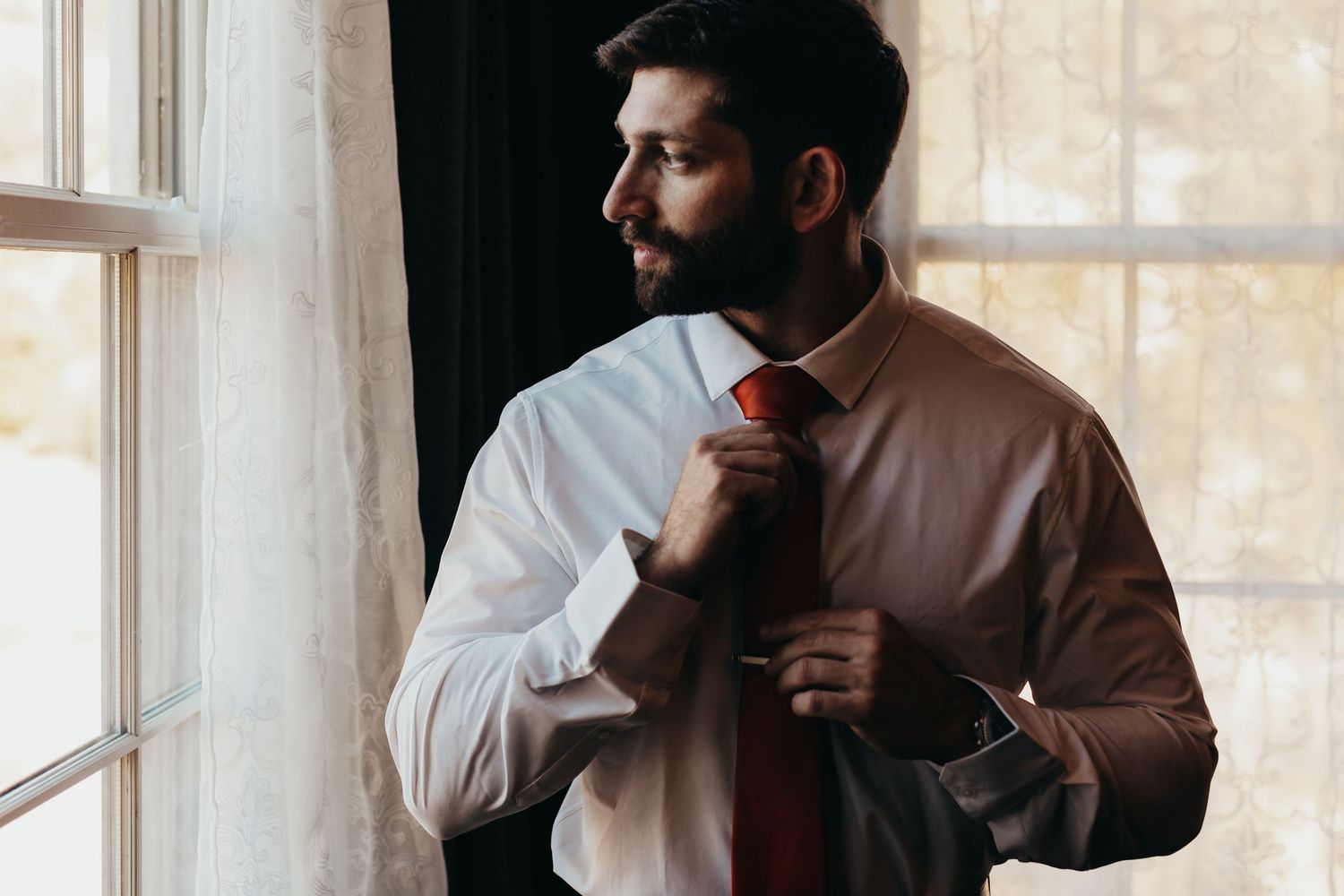Groom adjusts red tie while getting ready by window.